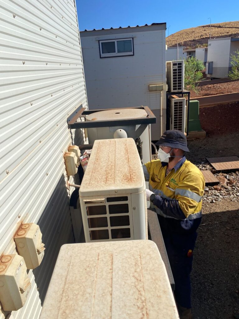 A worker in a yellow high-visibility jacket and hat, wearing a face mask, is repairing an outdoor air conditioning unit mounted on the side of a building. The area is dusty with several other AC units nearby.