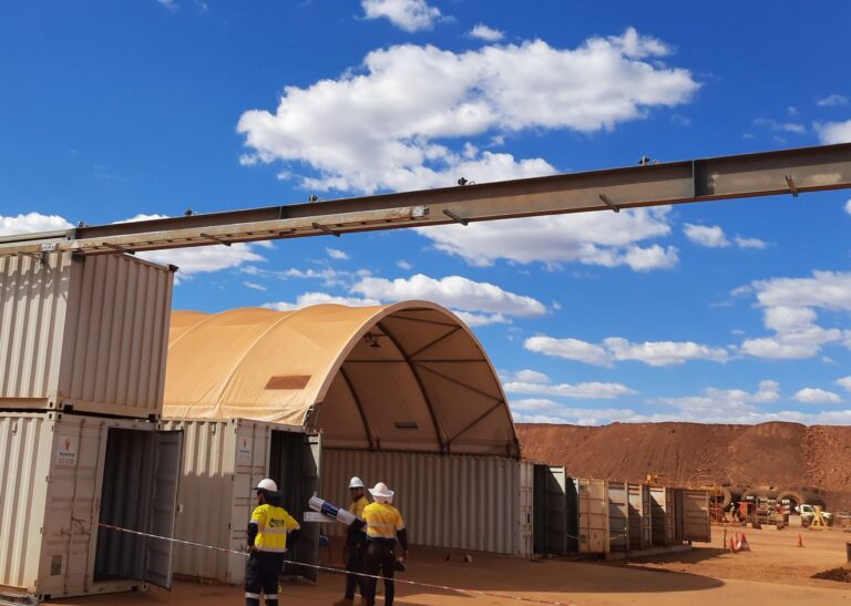 Construction site with workers in hard hats examining plans near stacked shipping containers. A large conveyor arm extends overhead. A curved fabric structure is visible in the background under a bright blue sky with scattered clouds.