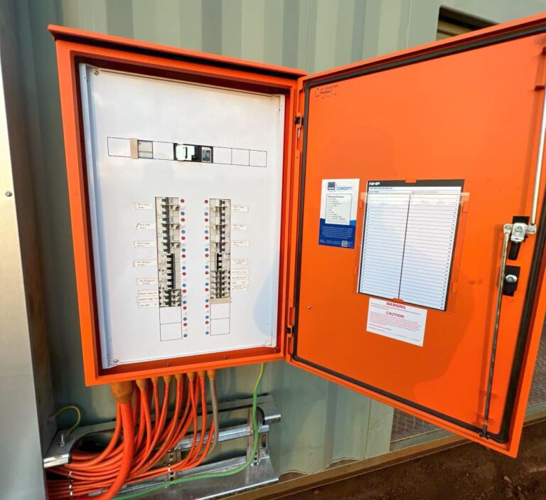 An open orange electrical panel with various switches and labeled circuit breakers. Several orange cables are connected at the bottom. The panel door has warning labels and a document holder with a clipboard. It's mounted on a metal structure.