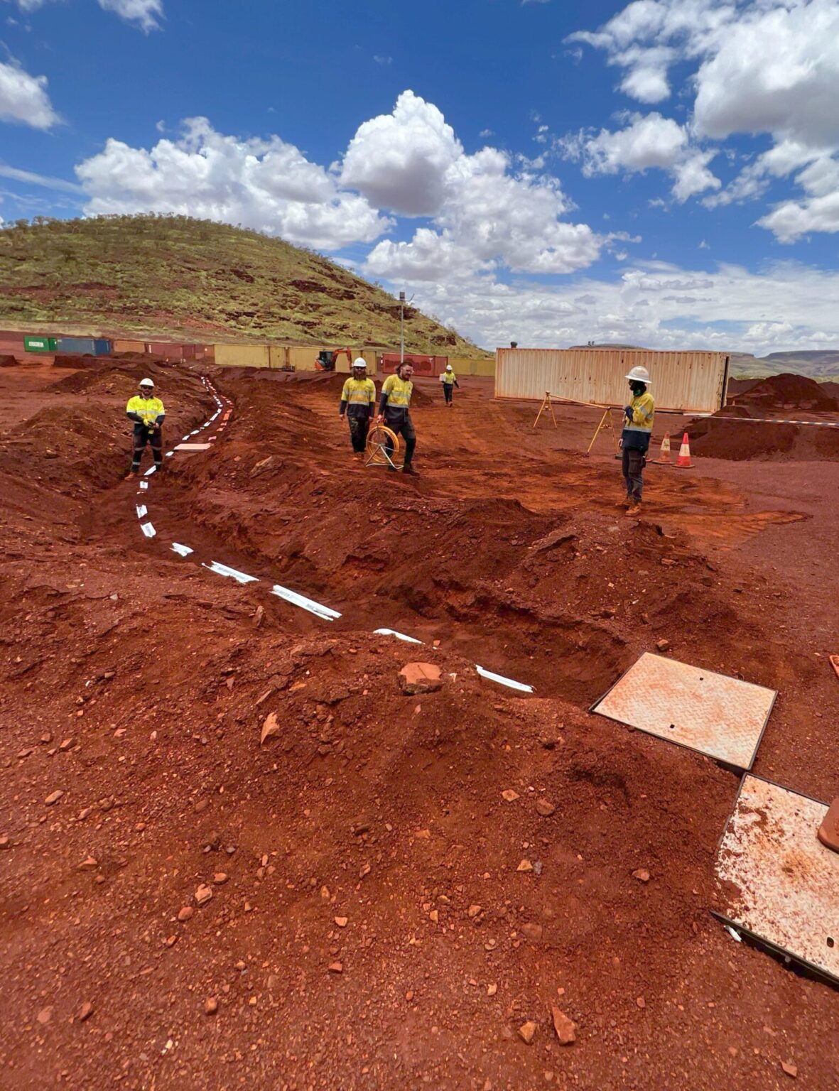 Workers in high-visibility clothing stand near a marked trench on a red dirt construction site, illustrating electrical HVAC and infrastructure solutions. Cones line the edge, hills rise in the background, and text overlay shares coordinates and project details.
