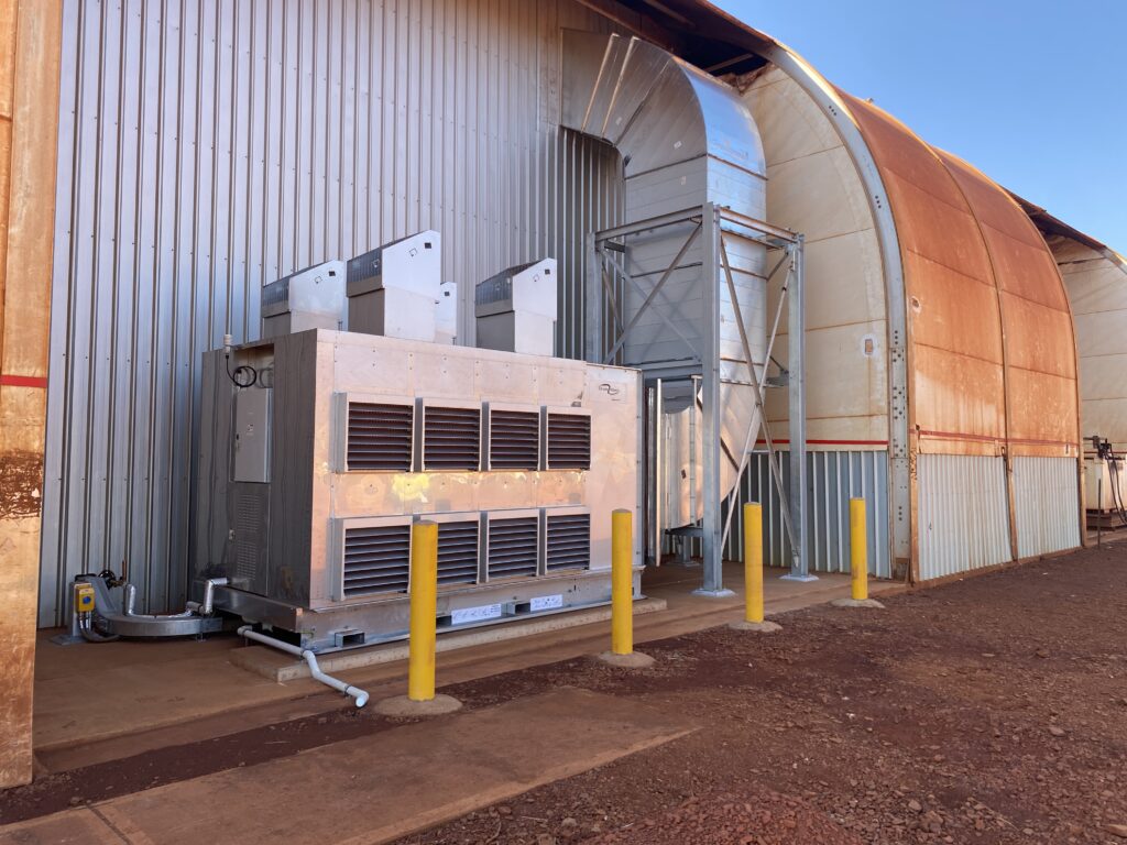 A large industrial air conditioning or ventilation unit with three exhaust vents is installed outside a corrugated metal building, protected by yellow bollards on a concrete pad, under clear blue skies.