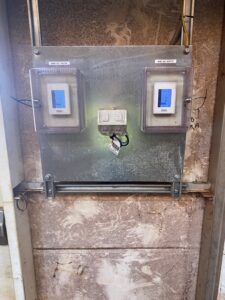 A metal panel mounted on a wall holds two enclosed digital displays, each labeled "AHU-11 SOUTH," flanking a central electrical outlet with plugged-in cords, all surrounded by a worn brown wall surface.