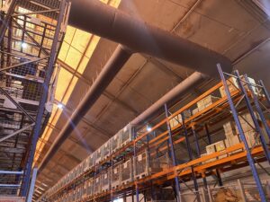 Large air ducts run along the ceiling of a warehouse with tall metal shelving units filled with boxes and crates. Warm light filters through the translucent ceiling panels.